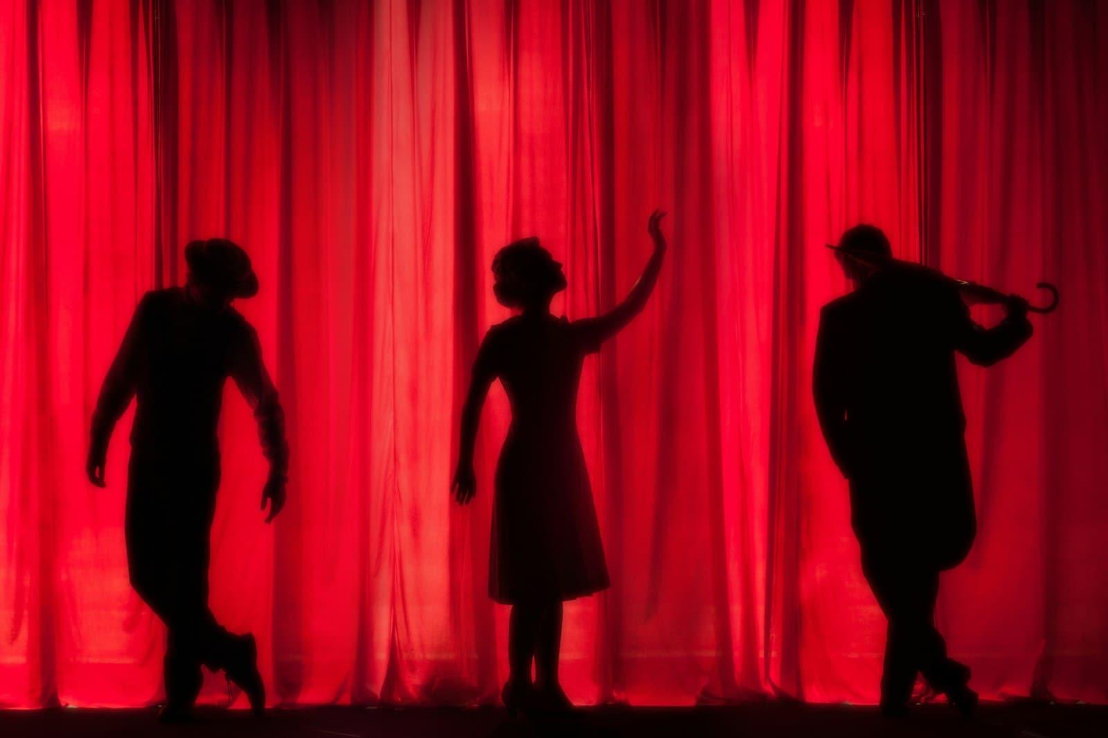 Siluetas de tres artistas frente a un telón rojo de teatro